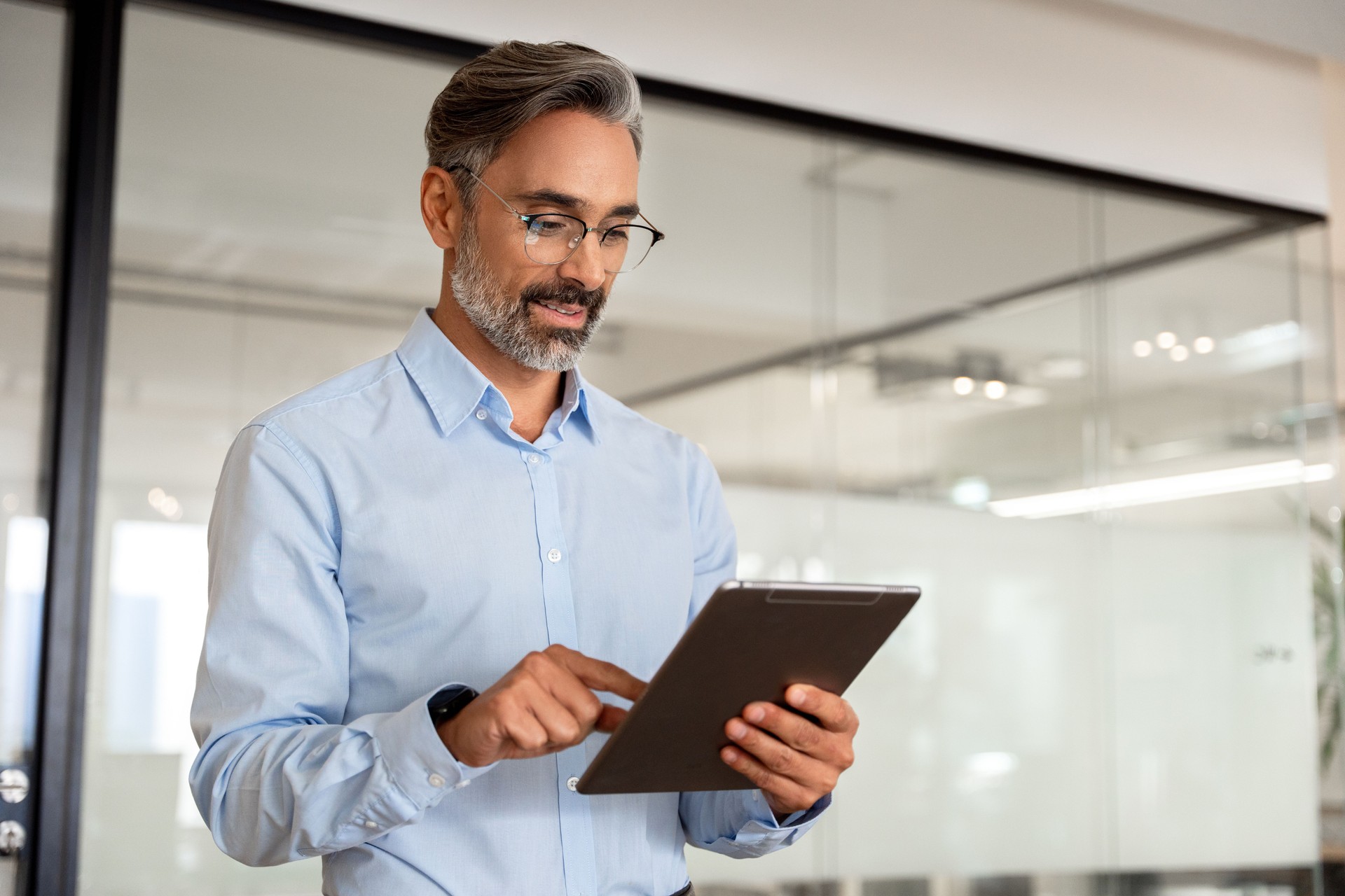 Happy Latin Hispanic bearded stylish mature adult professional business man, smiling Indian senior businessman CEO holding digital tablet using fintech tab application standing inside company office.